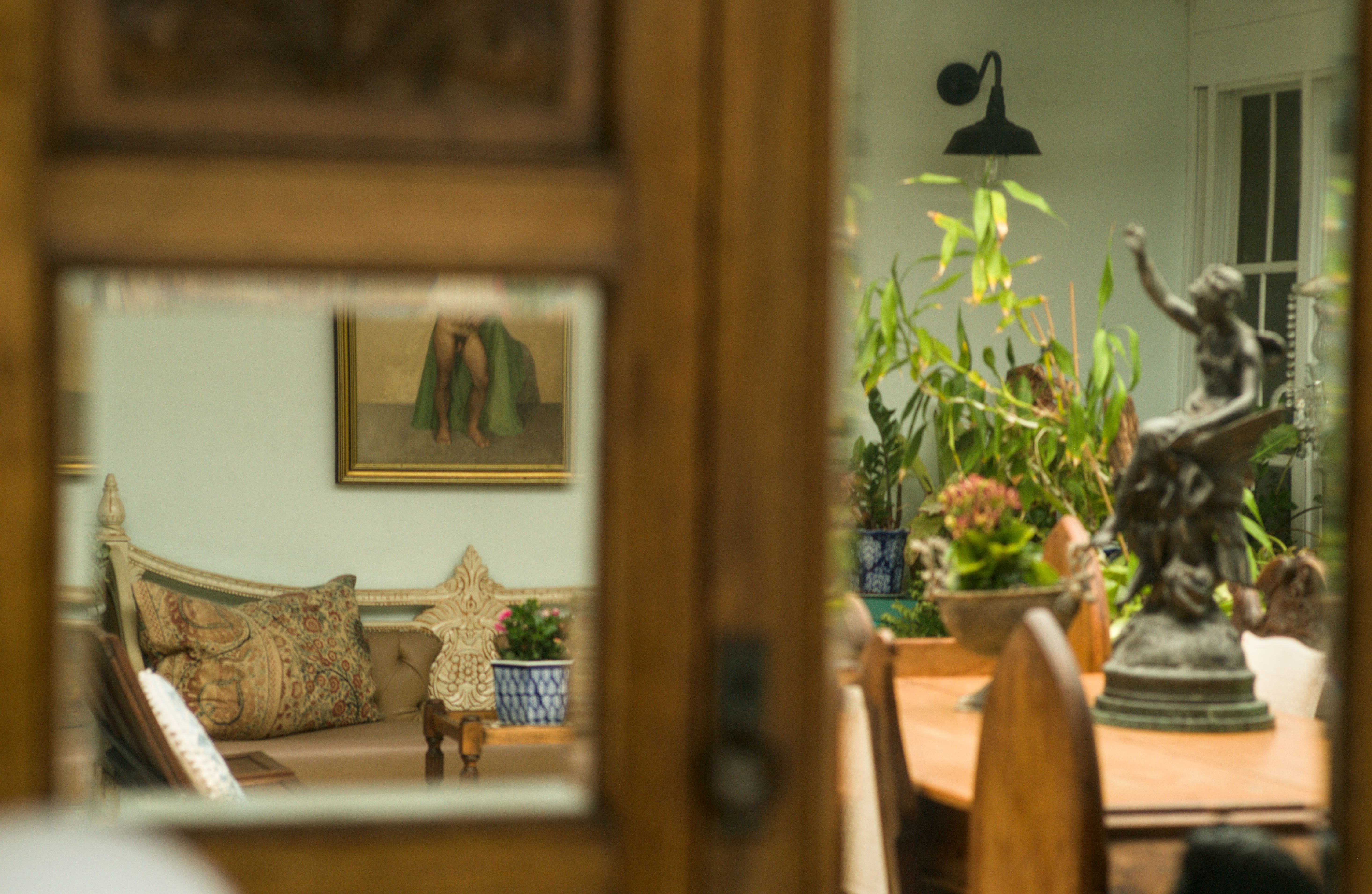Beauty Everyday, 1: Wide view of a garden room reflected in a cabinet mirror, with a sofa, plants, and a framed nude painting on the wall, lit by soft ambient daylight.