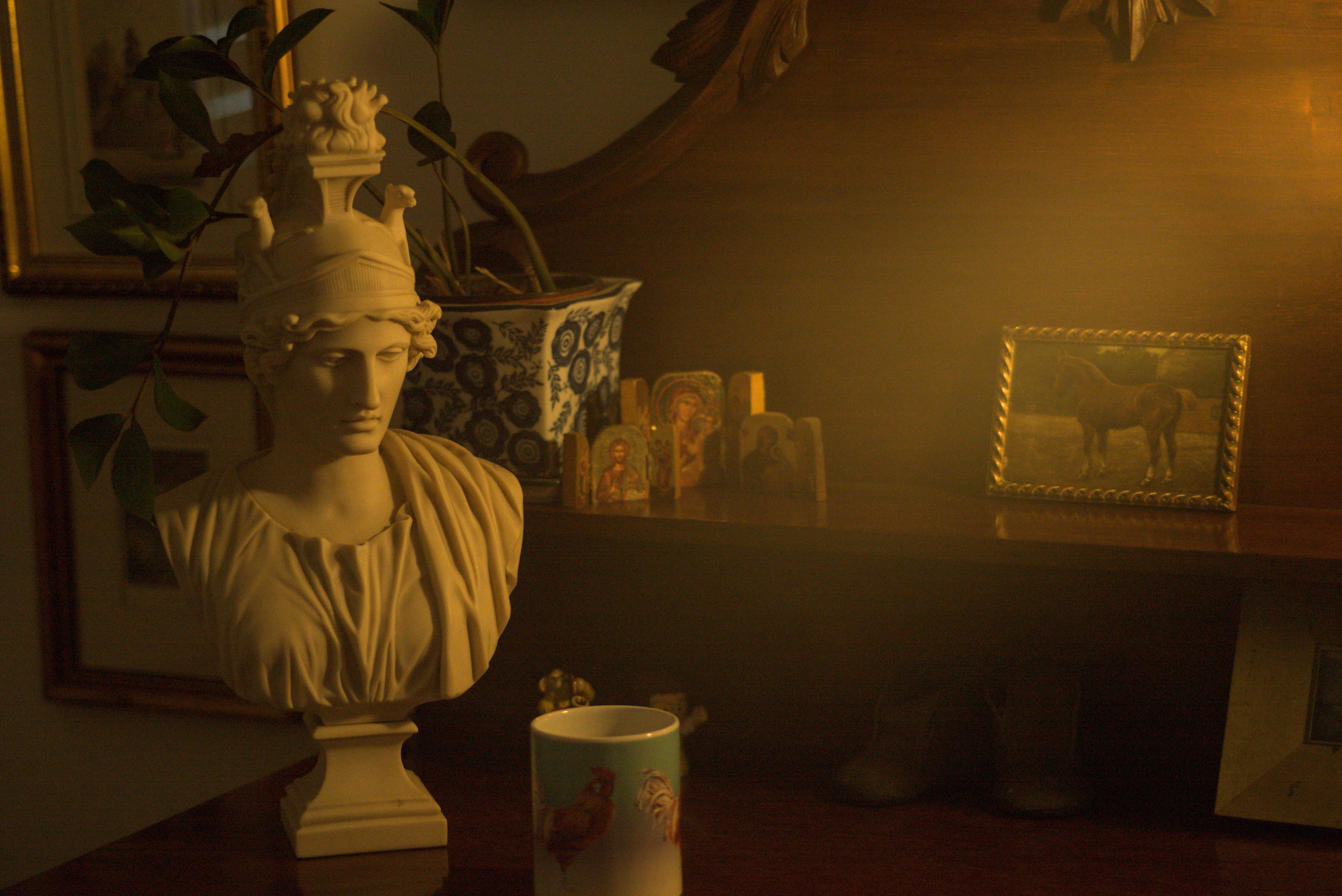 Beauty Everyday, 13: Detail of a lamp-lit tabletop holding a sculpture and personal items in a dim room.