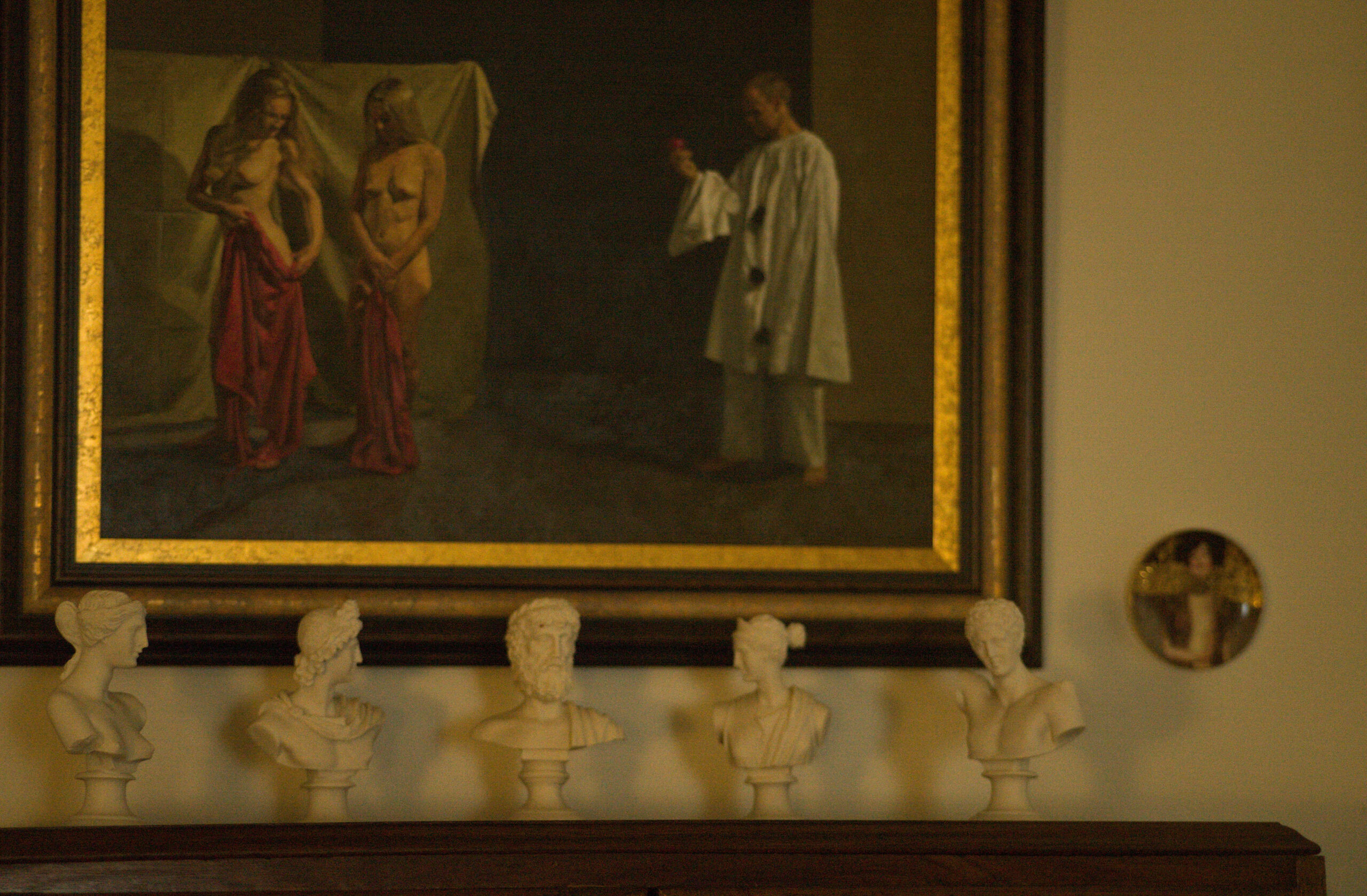 Beauty Everyday, 19: A series of small classical sculpted busts placed on a cabinet beneath a framed nude painting, lit by warm indoor light.