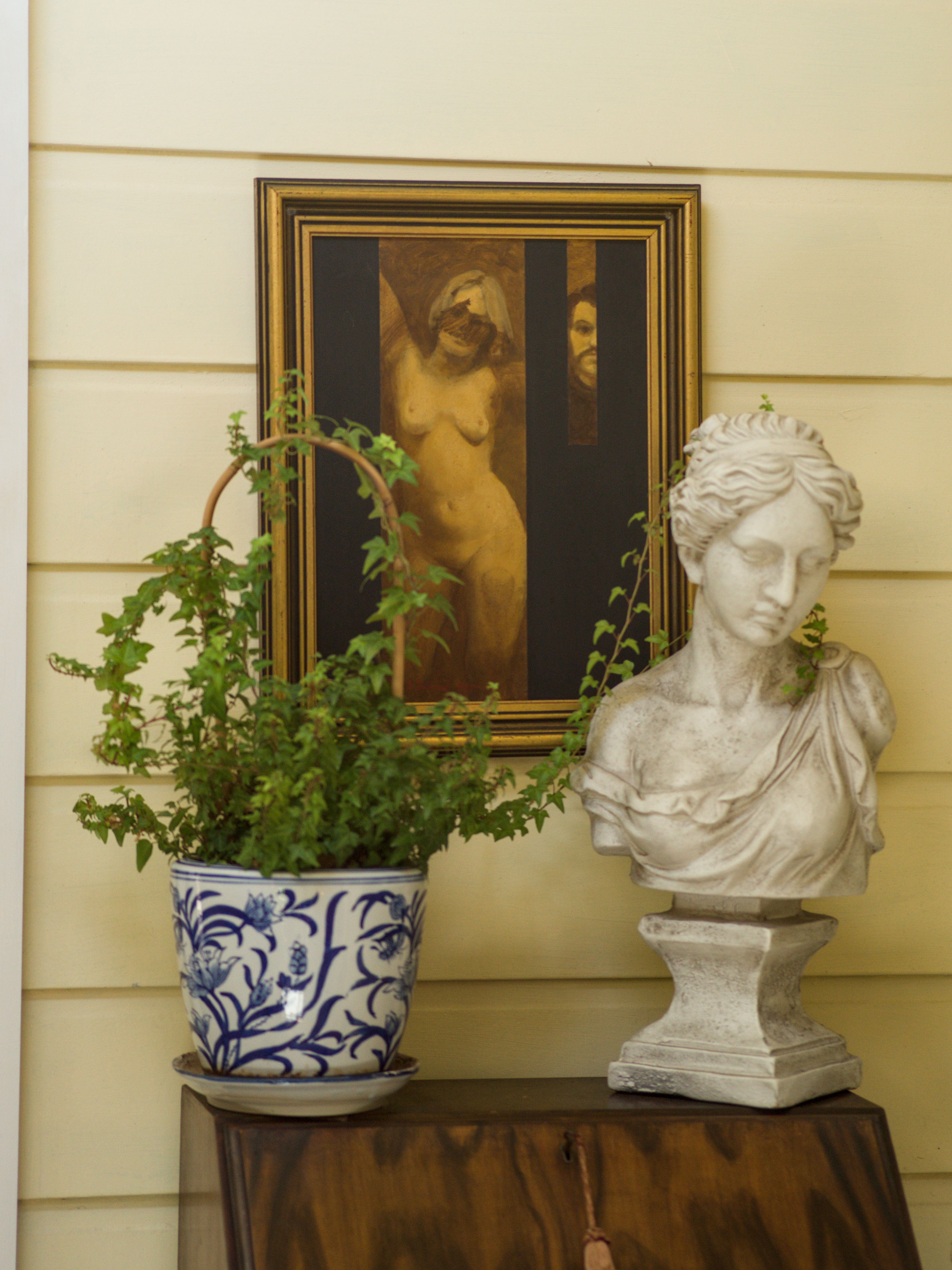 Beauty Everyday, 5: Classical sculpted bust on a cabinet beside a plant and a framed female nude, under neutral interior light.