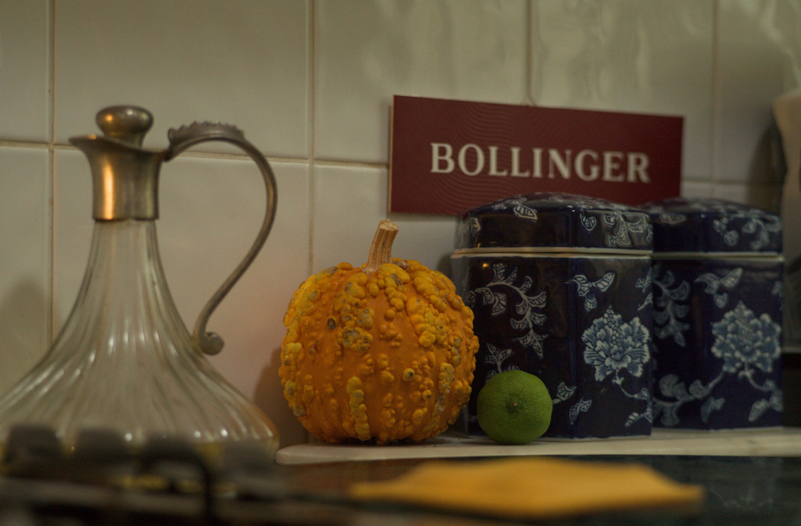 Beauty Everyday, 7: Kitchen area with jars, decanter, a pumpkin and a lime under dim interior light.