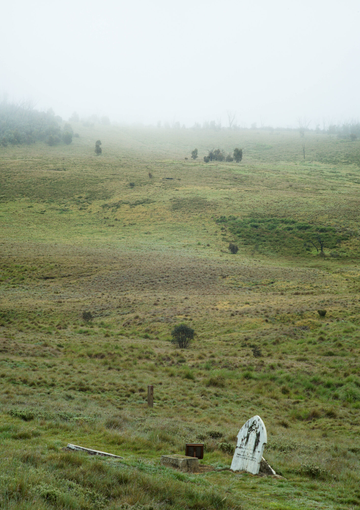 Remains, 6: A single remaining upright headstone at the base of a grassy hill obscured by morning fog.